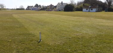 A grass field with a small post in the foreground and buildings in the background.