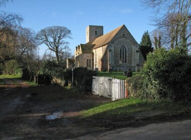 A stone church with a square tower seen across a small graveyard and wooden gate.