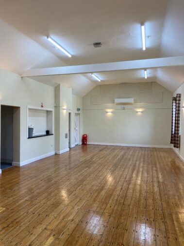 An empty hall with a high ceiling, wooden floors, and wall-mounted lights.