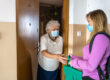 A young woman in a mask hands a green bag to an older woman in a mask at a door.