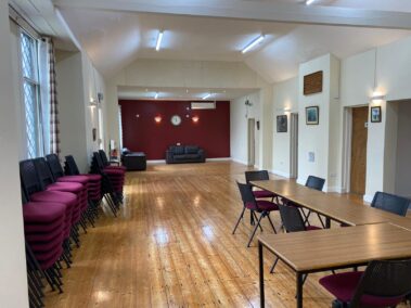 Large community hall with stacked chairs, wooden tables, and a red feature wall with a settee.