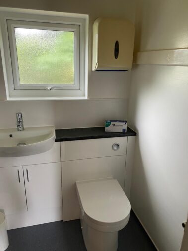 A bathroom with a white washbasin, toilet, and a wall-mounted paper towel dispenser.