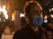 A person with hair in a bun wearing a blue face mask at night with blurred city lights.