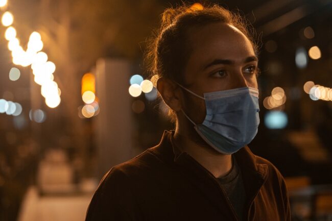 A person with hair in a bun wearing a blue face mask at night with blurred city lights.