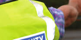 Person in a yellow Community Speedwatch vest holds a speed gun at the roadside.