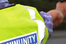 Person in a yellow Community Speedwatch vest holds a speed gun at the roadside.