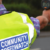 Person in a yellow Community Speedwatch vest holds a speed gun at the roadside.