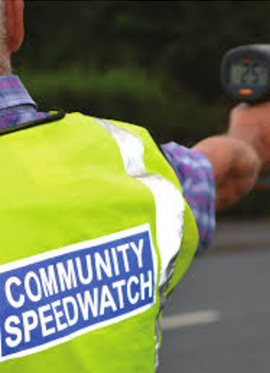 Person in a yellow Community Speedwatch vest holds a speed gun at the roadside.