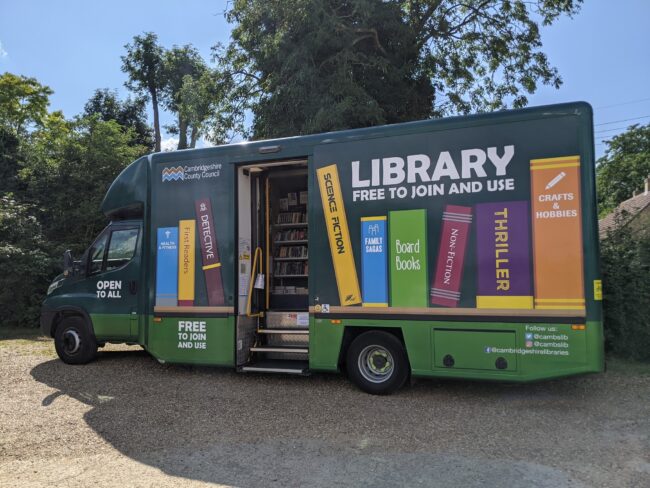 Green mobile library van with books painted on the side, parked outdoors with door open. Green mobile library van with books painted on the side, parked outdoors with door open.